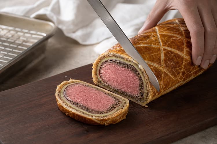 Beef Wellington method step 8, Rest to 63°C, carve, and sauce beside: The slice shows even pink beef, thin distinct mushroom, ham, and green crêpe layers, a dry crisp base, and almost no juice on the board.